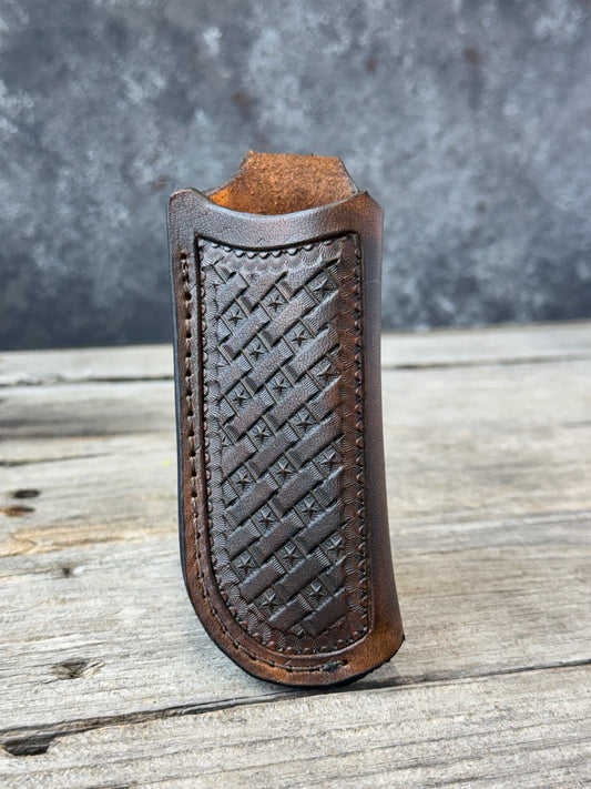 A close-up of a dark brown custom leather knife sheath with a woven basketweave pattern on the front, resting on a weathered gray wooden surface.