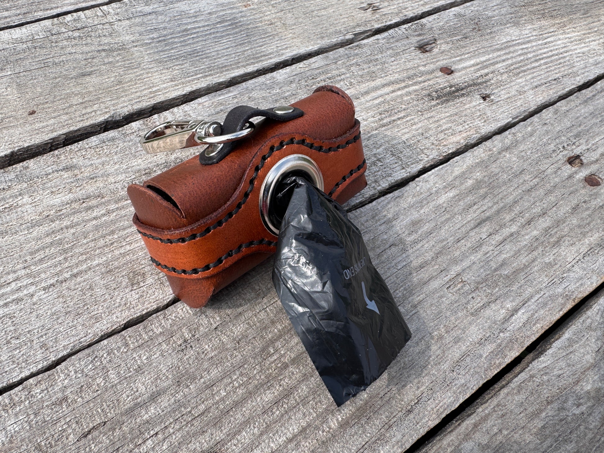 A brown leather dog poop bag holder keychain with a black plastic bag pulled out, resting on weathered gray wooden planks.