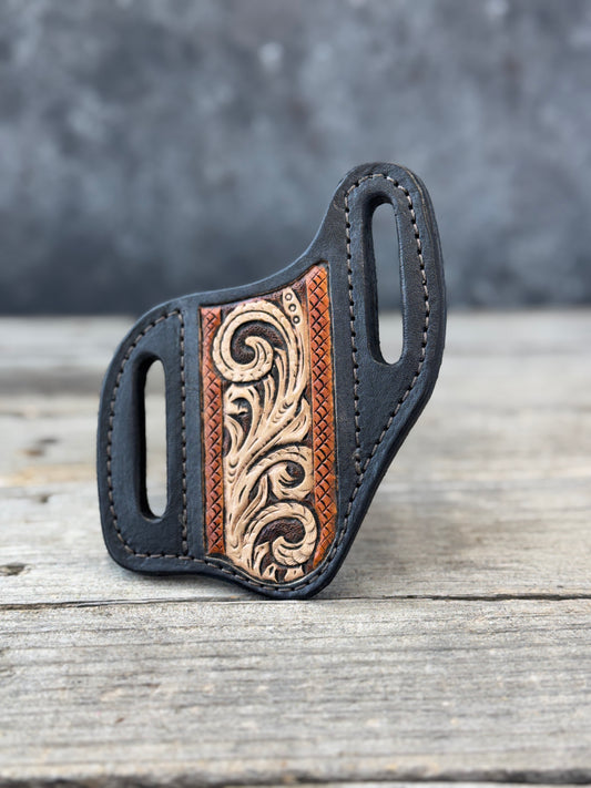 A black custom leather knife sheath with a carved brown and beige floral leather panel, resting on a rustic gray wooden surface.